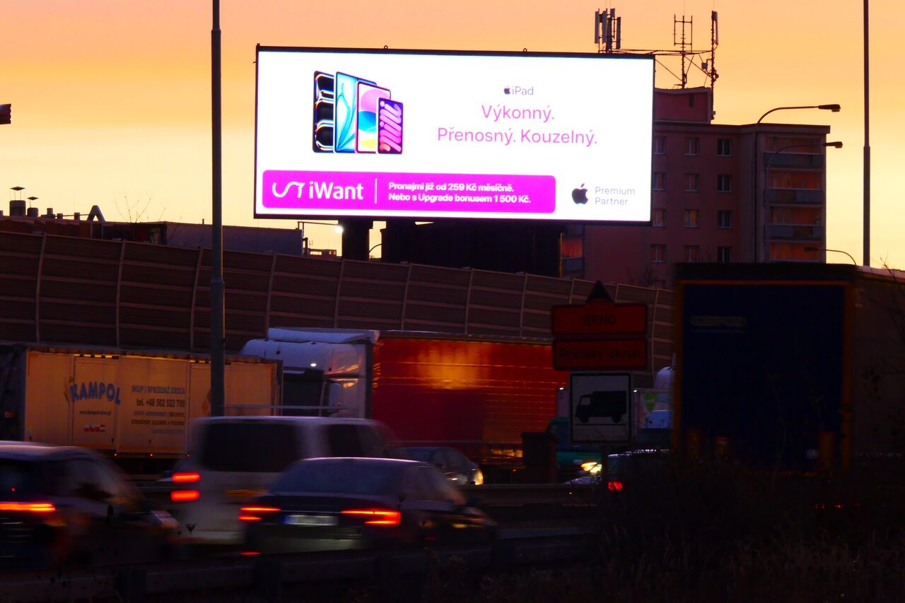 Velkoplošná LED obrazovka s kampaní iWant nad rušnou výpadovkou při západu slunce