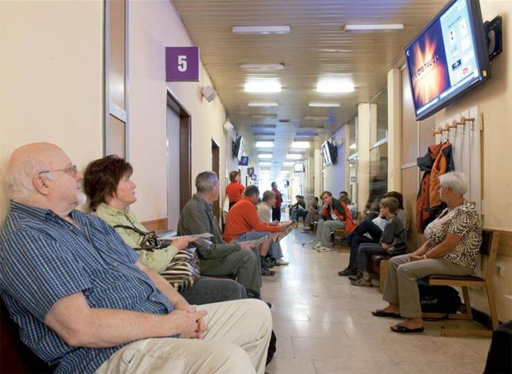 Čekárna zdravotnického zařízení: lidé sedí na lavicích a na stěně nad nimi visí TV obrazovka s vysíláním